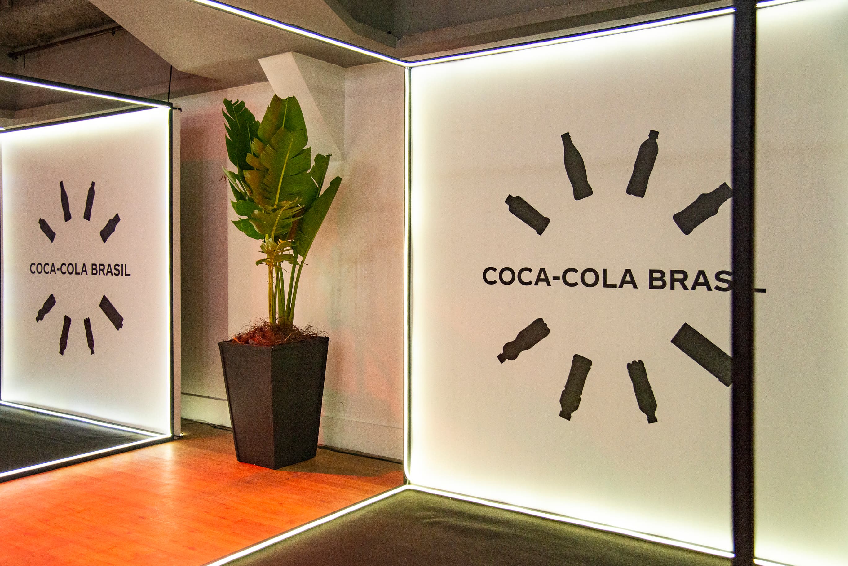 Parede de branding "Coca-Cola Brasil" com planta decorativa em ambiente de evento