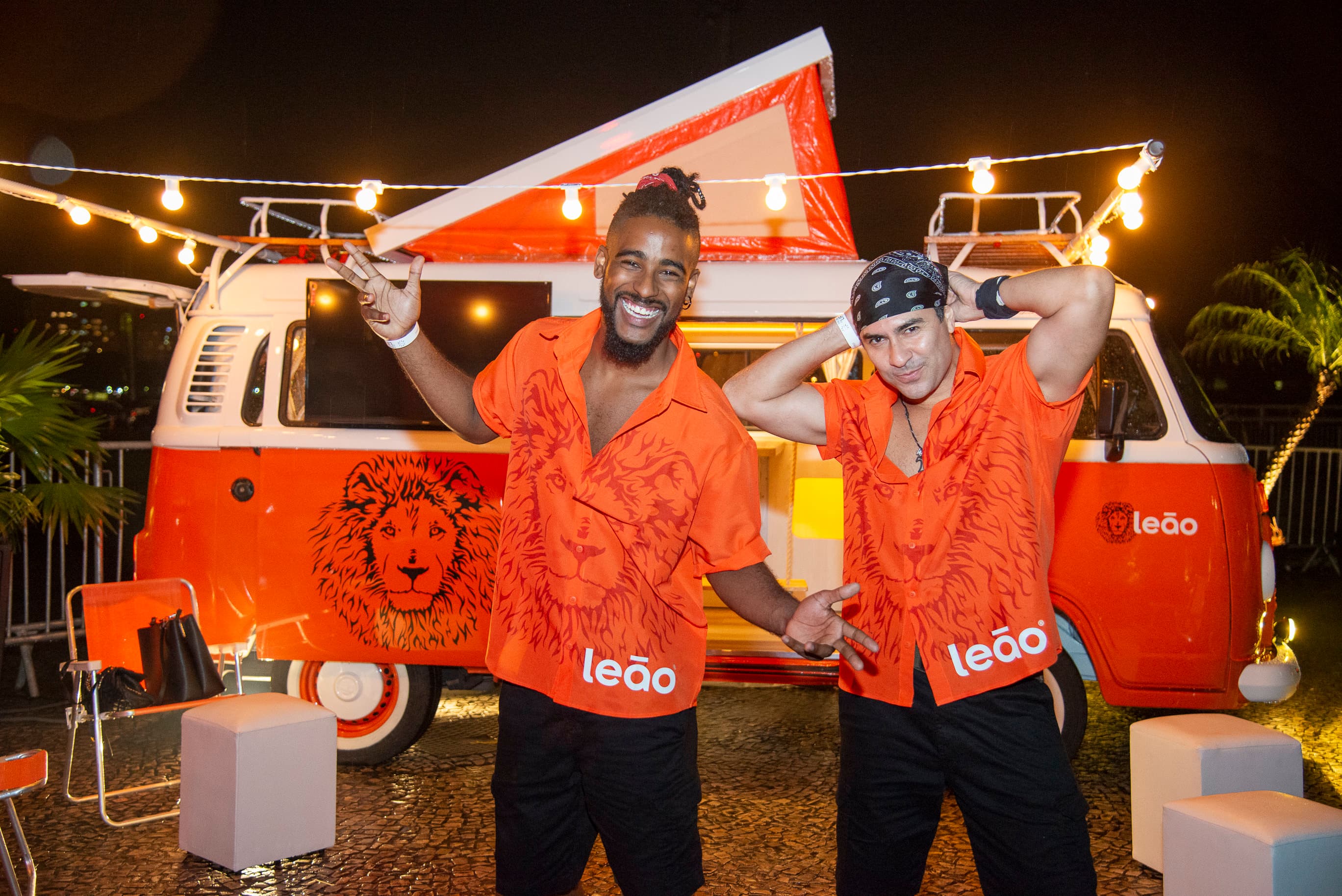 Dois participantes de camisa laranja "Leão" posando ao lado de kombi vintage em evento
